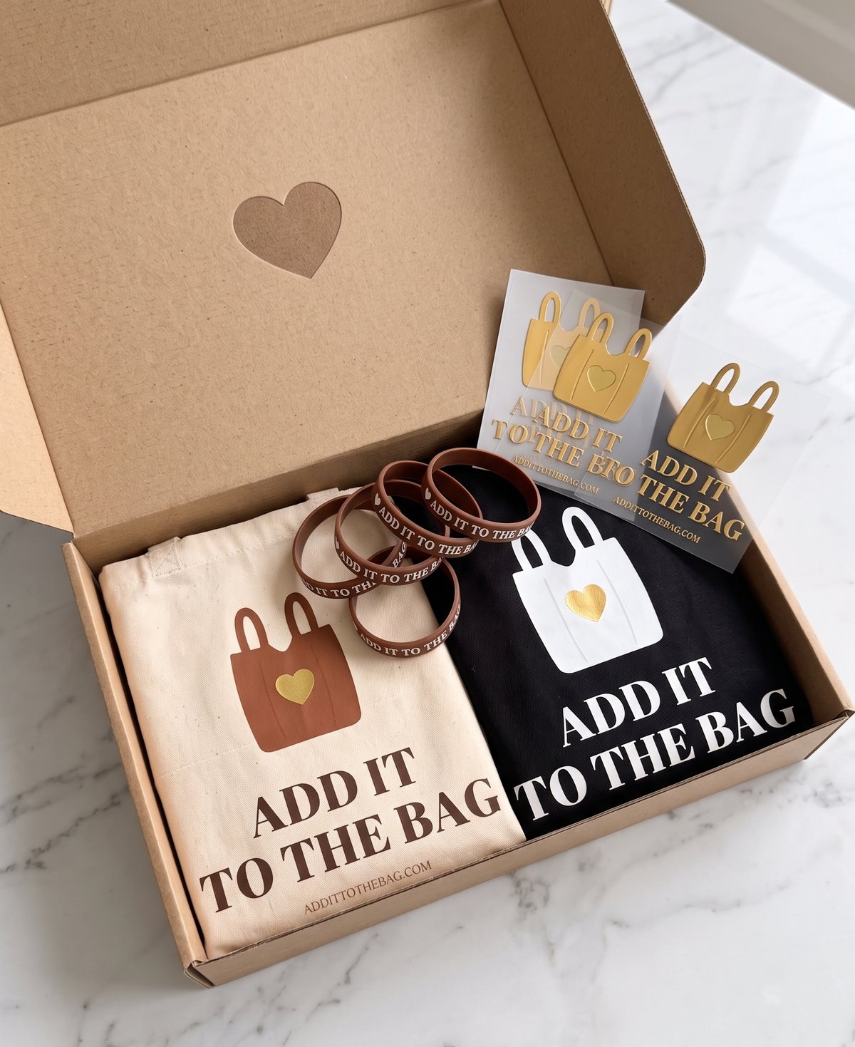 The Add It To The Bag box: two canvas totes (one natural, one black), a stack of wristbands, and gold logo stickers packed in a keepsake heart box